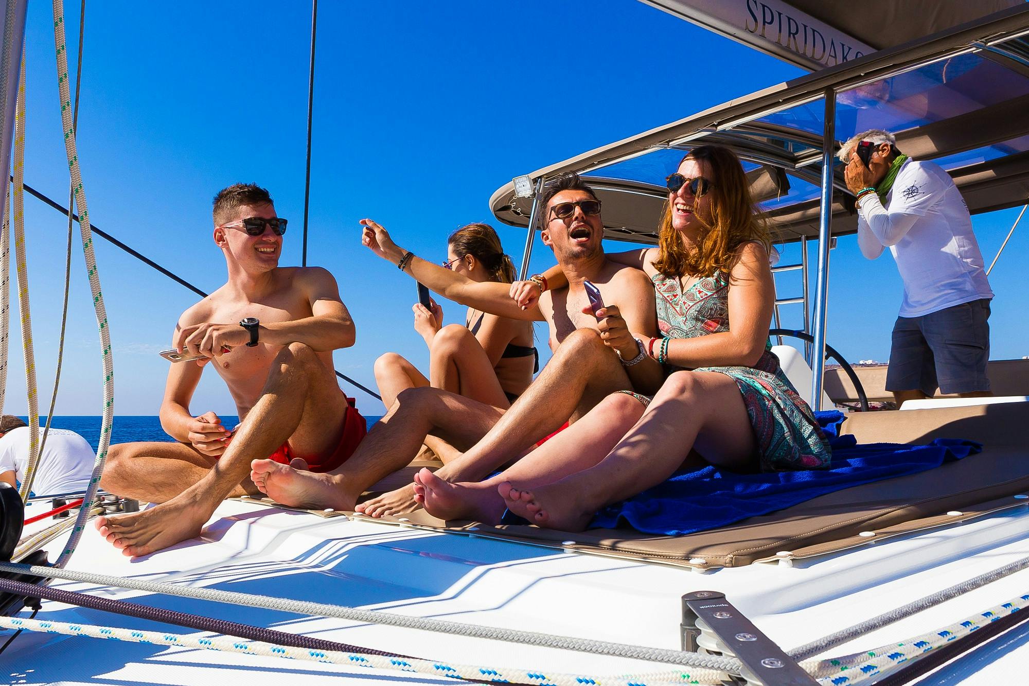Un gruppo di giovani adulti si rilassa e ride sul ponte di una barca a vela in un cielo azzurro e limpido. Una persona regola le vele.