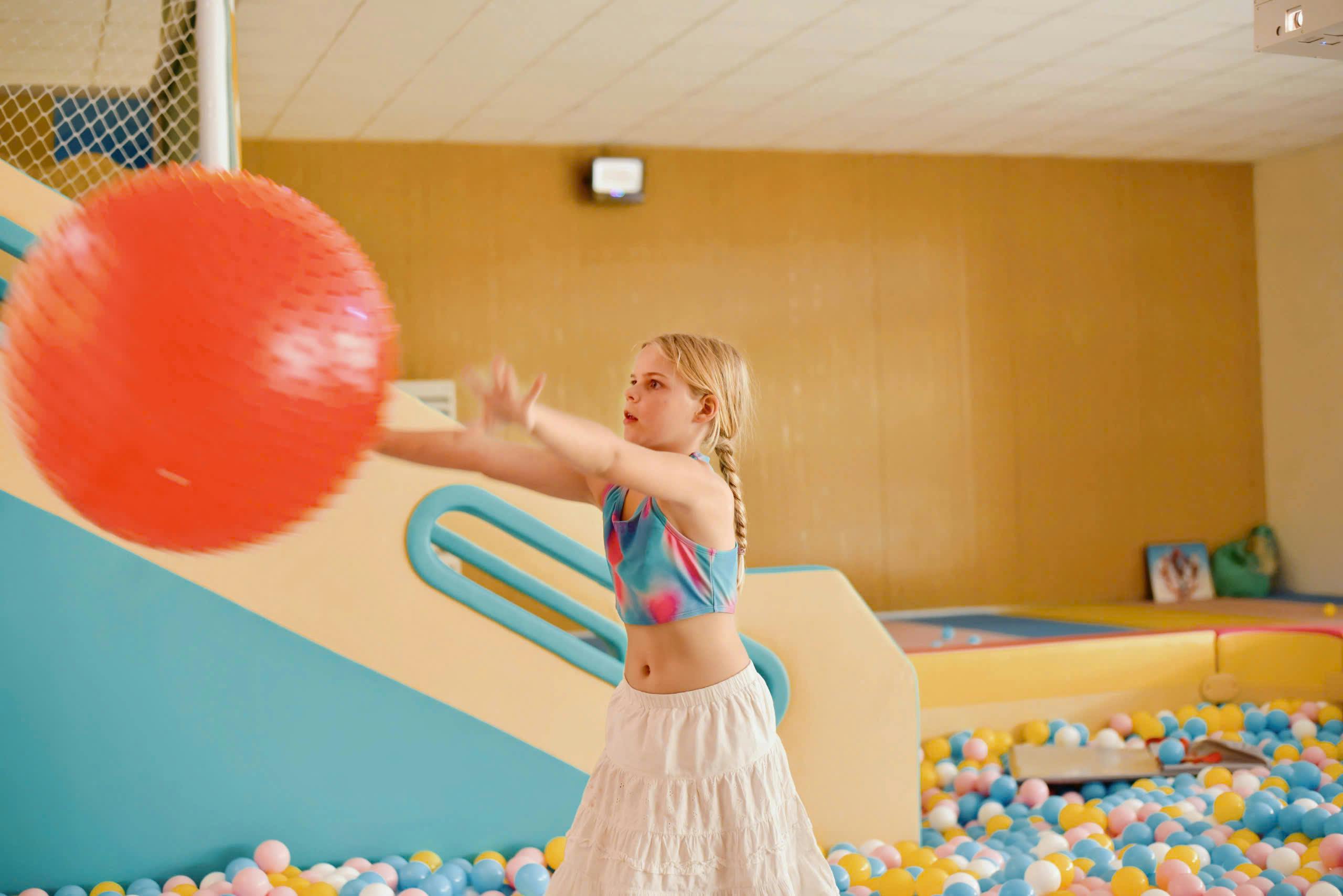 A girl in a colorful top and white skirt plays with a large red ball in an indoor play area with a ball pit and slide.