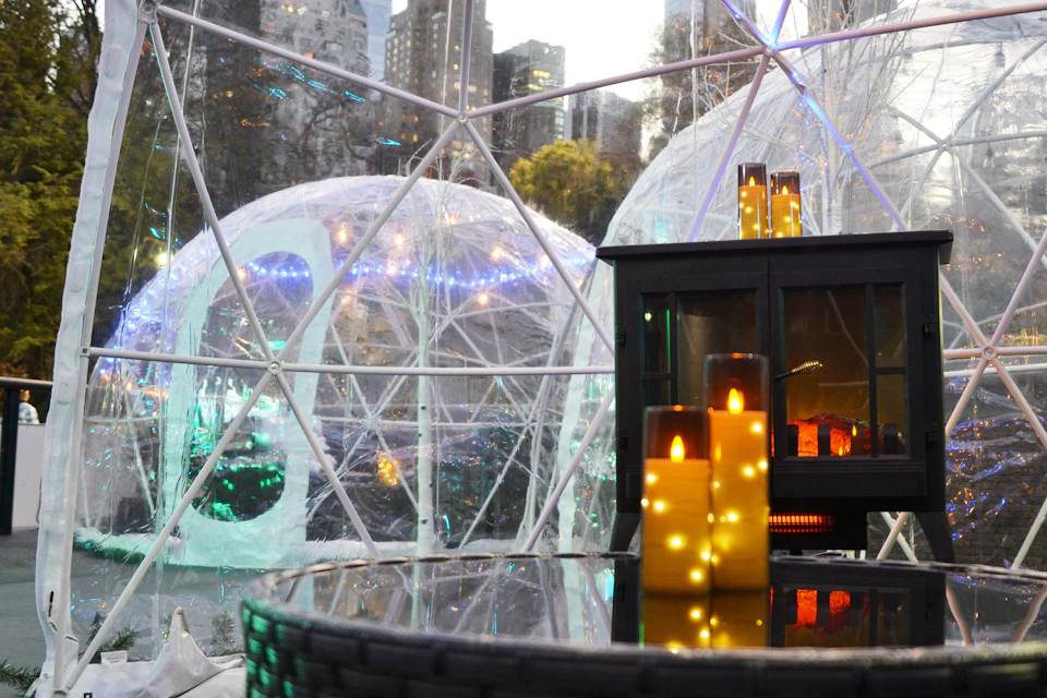 Geodesic domes with fairy lights and glowing candles on a nearby table. Cityscape and trees visible outside.