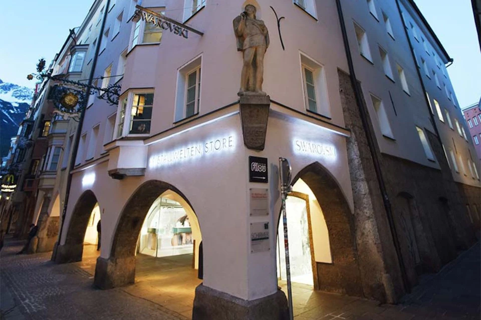 Illuminated Swarovski store exterior with arched doorways, large windows, and a statue on the corner of the building at dusk.