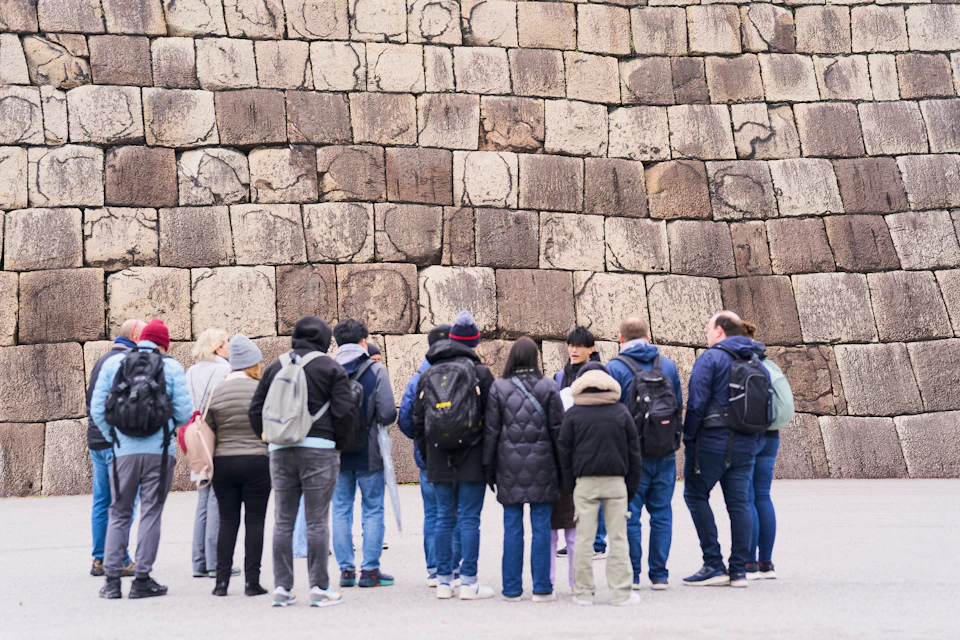 A group of people standing in front of a large stone wall, some wearing winter clothing and backpacks.