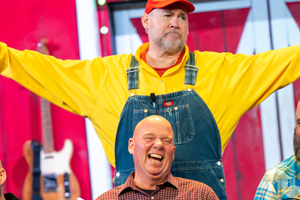 A performer in colorful attire with props stands behind three laughing seated men against a red barn backdrop.