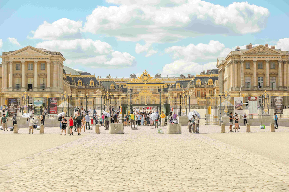 Tourists walking and gathering in front of the Palace of Versailles' golden gates on a sunny day.