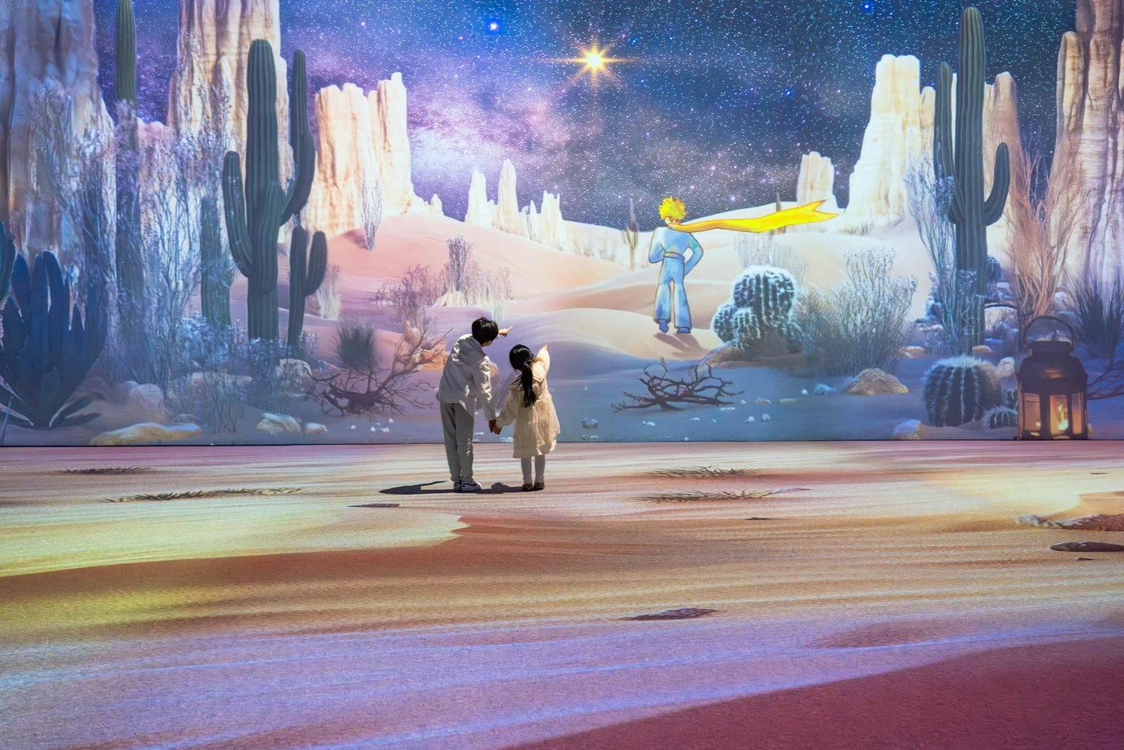 两个孩子手牵手站着，看着一个以沙漠为主题的投影，其中有动画人物小王子。