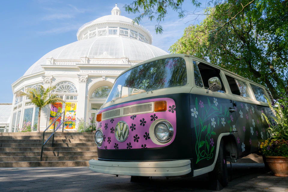 A vintage Volkswagen van with a floral design parked in front of a large domed conservatory building on a sunny day.