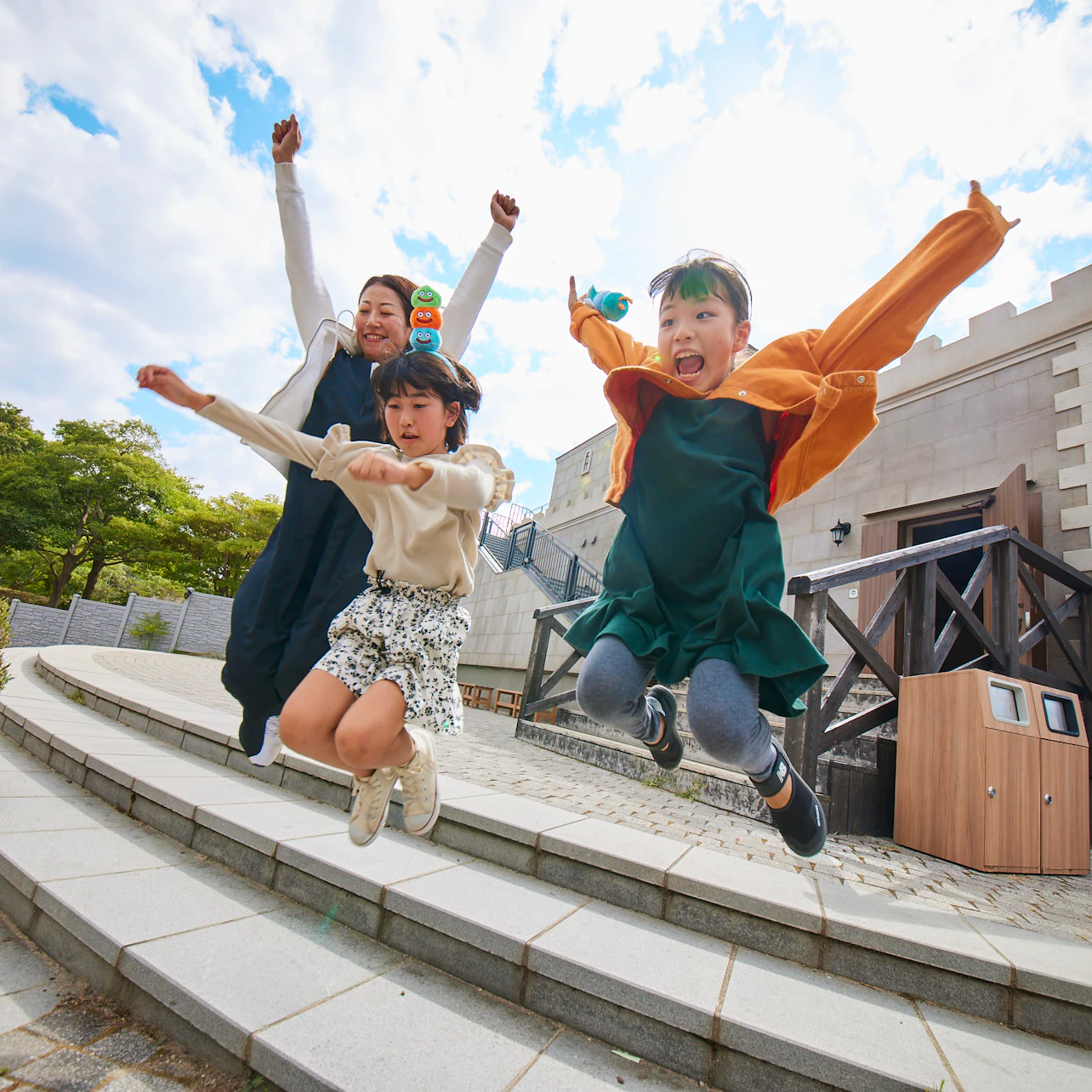 Three people, two children and one adult, jumping joyfully on steps outdoors in front of a building on a sunny day.