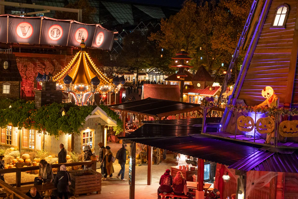 A nighttime festival scene with colorful lights, a carousel, people walking, and various stalls and decorations.