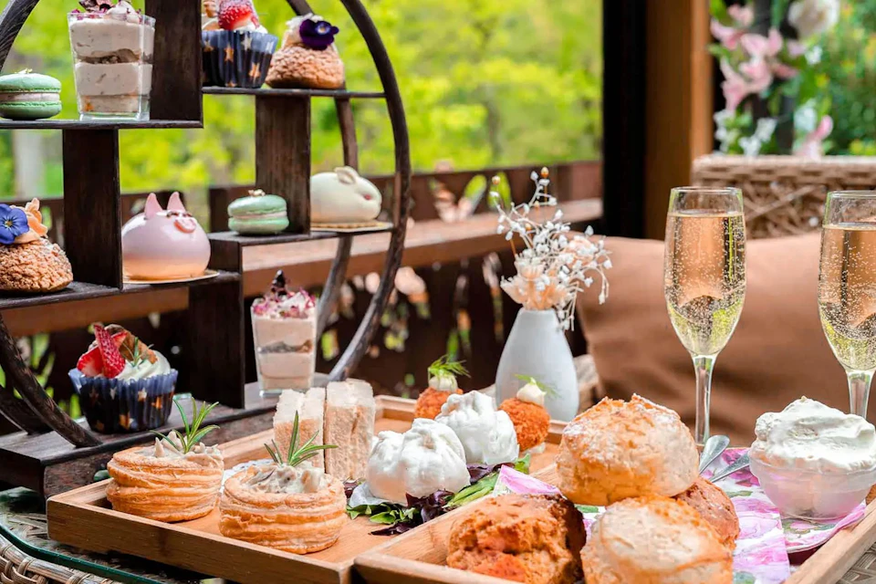 A wooden table set with tea, scones, pastries, and a glass of champagne, with a tiered stand of desserts in a cozy setting.