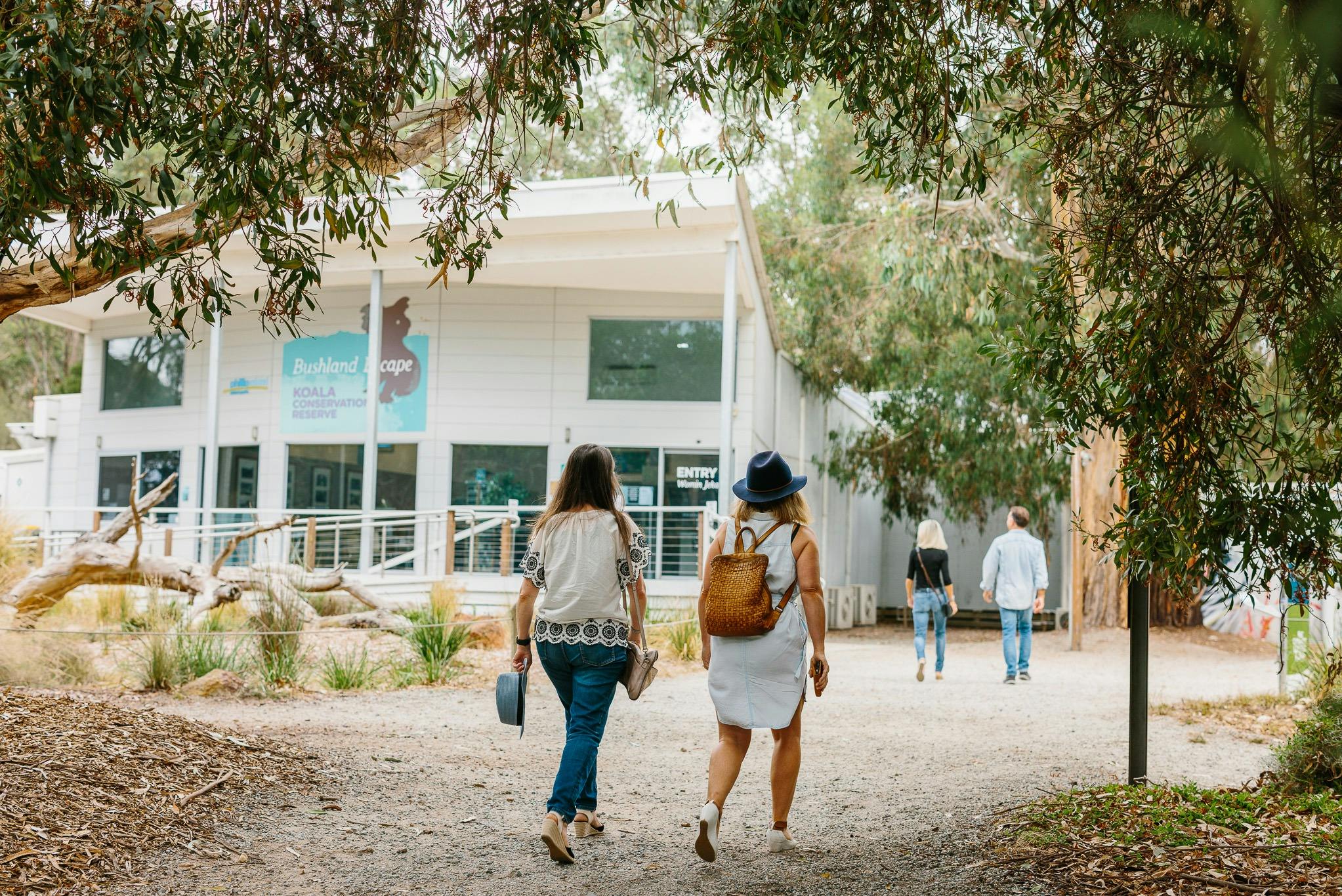 People walking towards a building surrounded by trees, with a sign reading "Bushland Escape Koala Adventure."