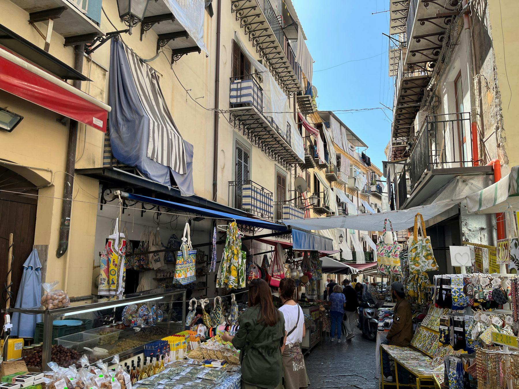 A bustling street market with stalls selling goods, including bags and food items, surrounded by buildings with balconies.