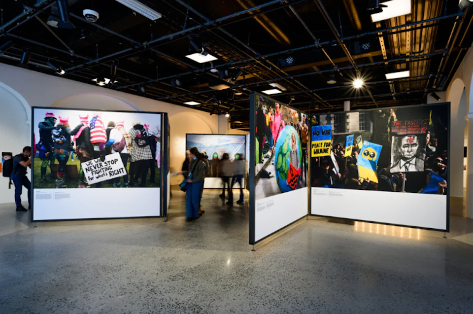 Indoor gallery with large photos of protests displayed on freestanding panels. People walk between the exhibits.