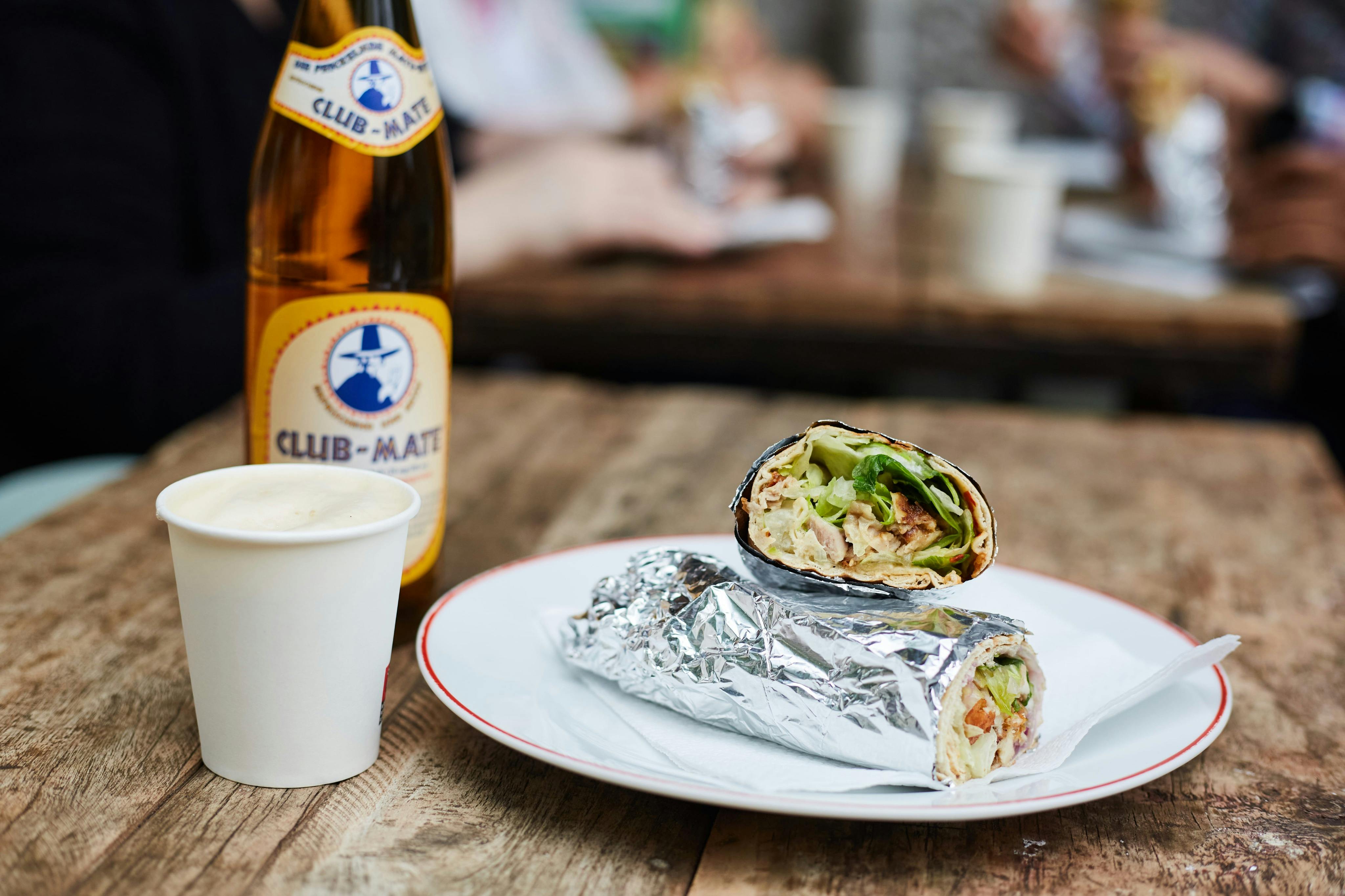 Ένα πιάτο με ένα μπουρίτο τυλιγμένο σε αλουμινόχαρτο και ένα κομμένο μισό, μαζί με ένα μπουκάλι Club-Mate και ένα φλιτζάνι σε ένα ξύλινο τραπέζι.