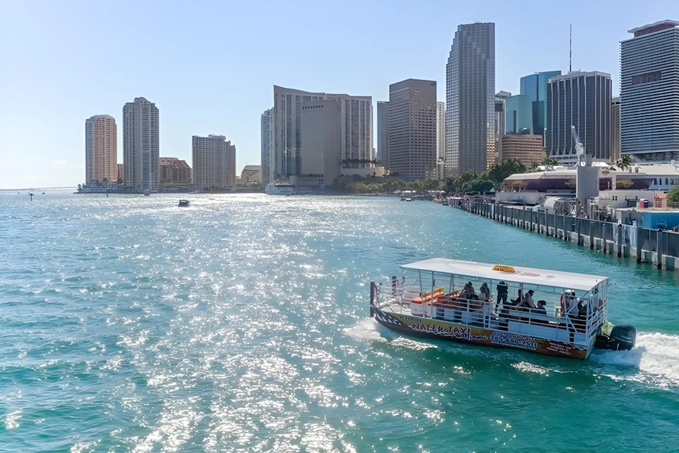 A water taxi is cruising on a blue waterway with a city skyline of tall buildings in the background under a clear sky.