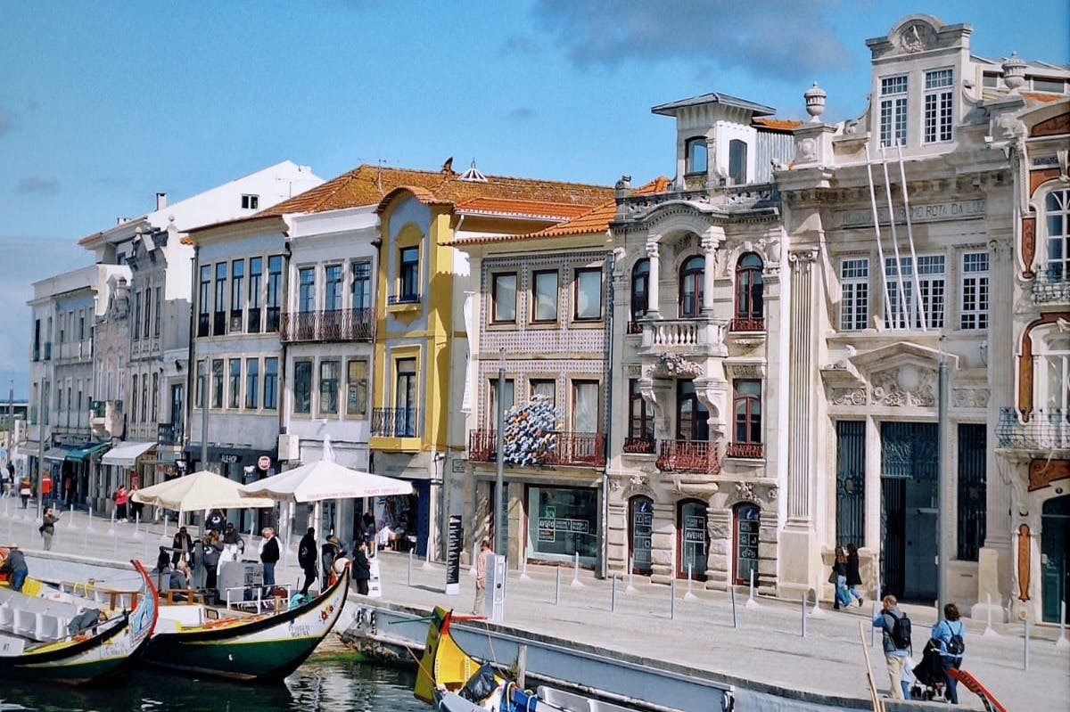 Πολύχρωμα, περίτεχνα κτίρια σχηματίζουν ένα κανάλι με βάρκες αγκυροβολημένες. Οι πεζοί περπατούν κατά μήκος της παραλίας κάτω από καθαρό ουρανό.