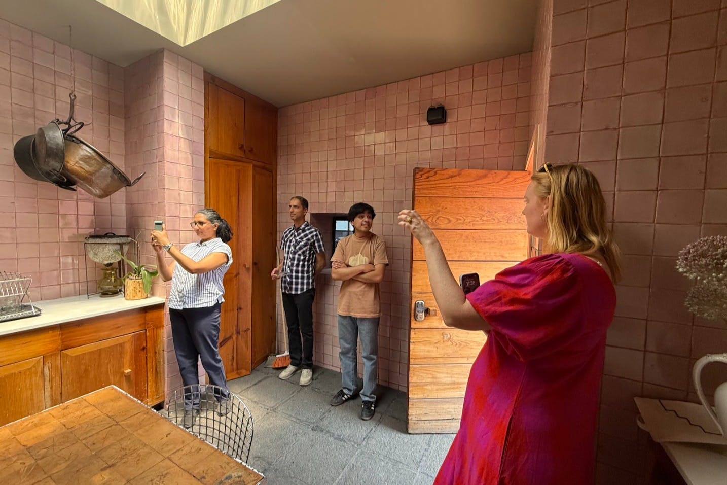 人们观察厨房，厨房里有粉色瓷砖墙和木制橱柜。 一个人用手机拍照。