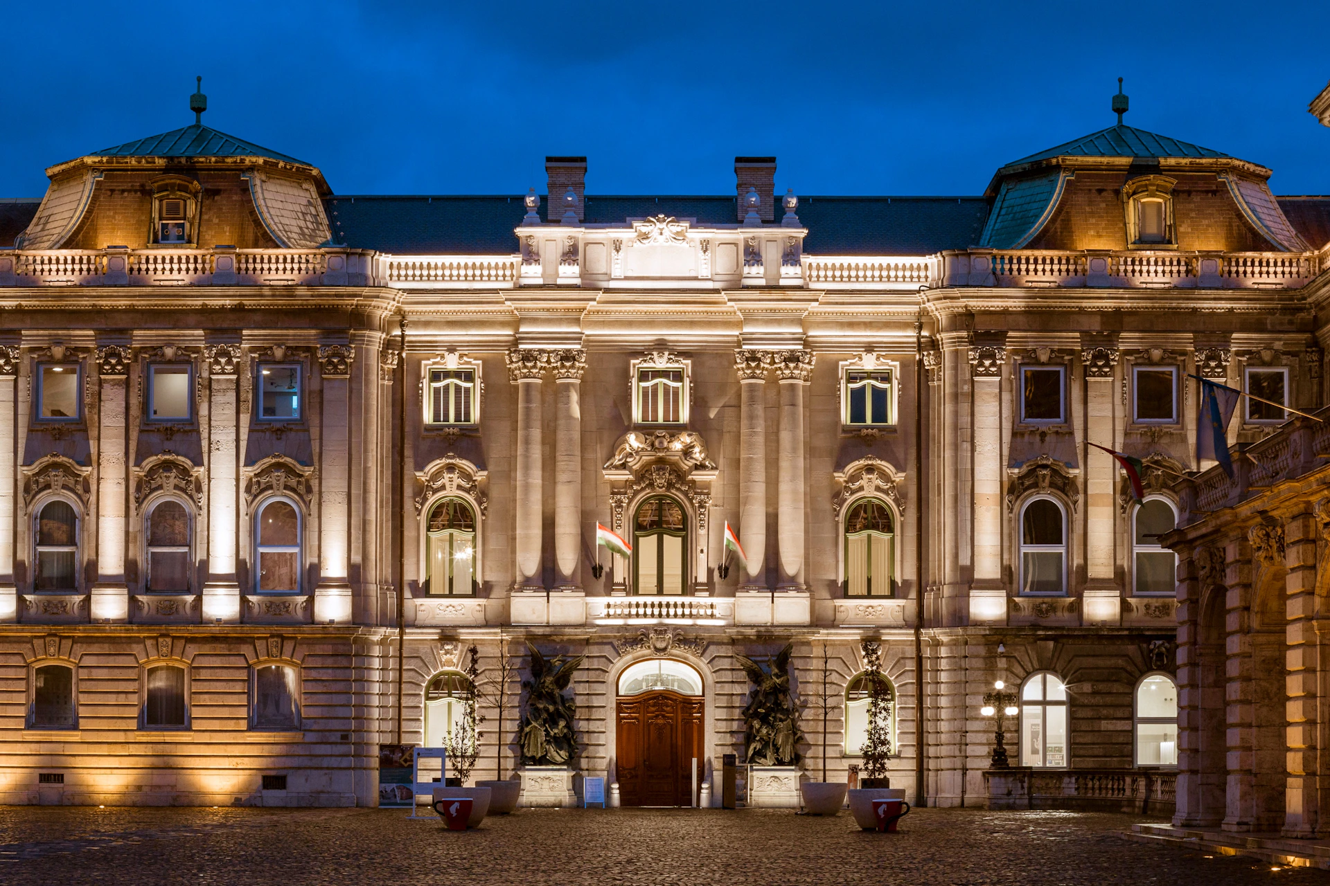 Illuminated historic building with ornate architectural details at night, featuring statues and flags by the main entrance.