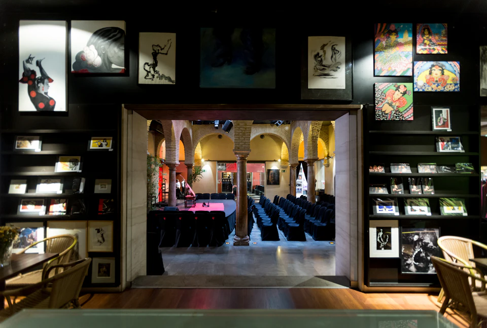 Room with arched ceilings and brick columns, set up with rows of black chairs facing a stage. Artwork is displayed on the surrounding walls.