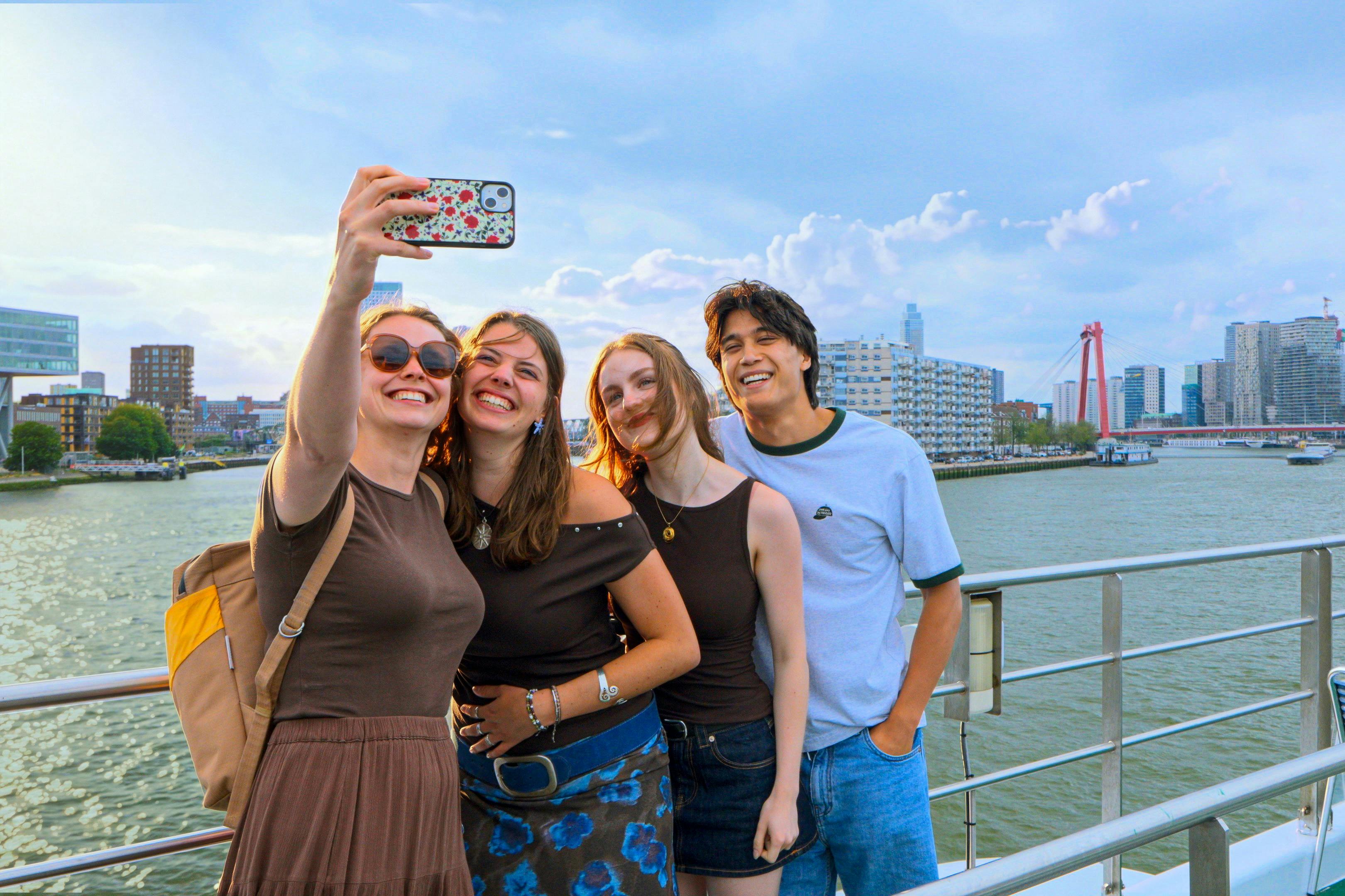 Un gruppo di quattro persone che sorridono e si scattano un selfie su una passeggiata sul lungomare, con edifici e acqua sullo sfondo.