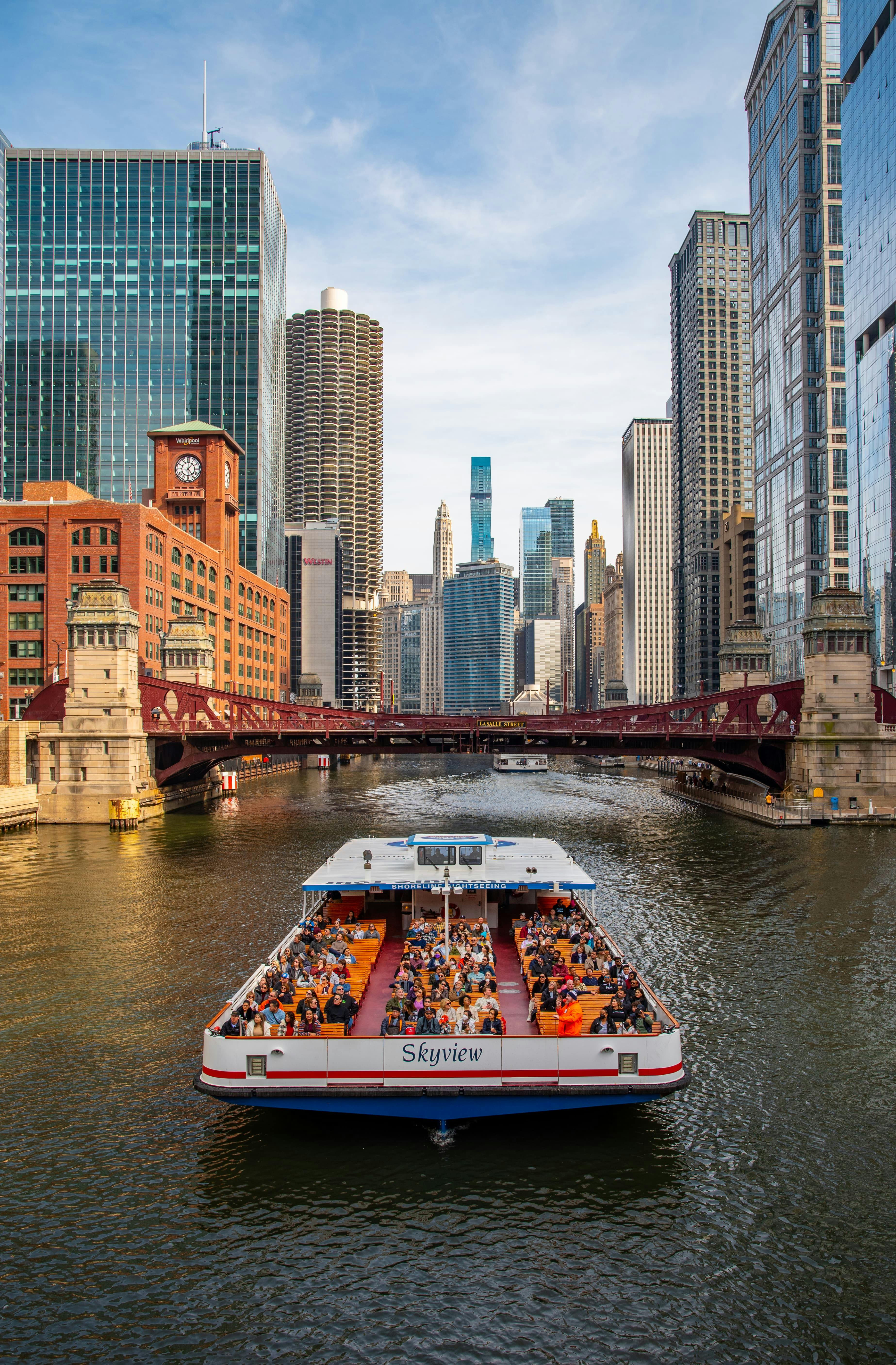 高層ビルとモダンな建築が並ぶ街で、人々でいっぱいのボートが赤い橋に近づいています。