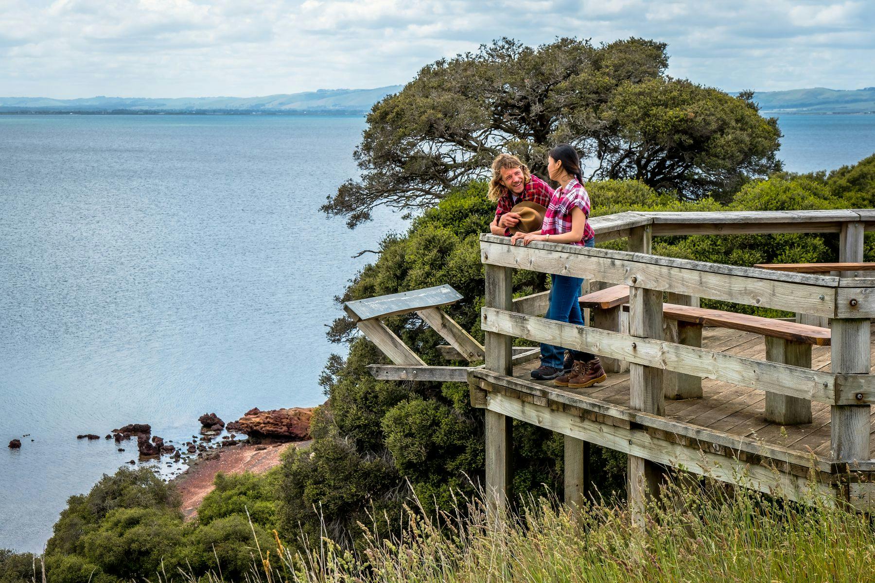 Deux personnes sur un belvédère en bois, surplombant un plan d'eau pittoresque avec des arbres et des falaises en arrière-plan.