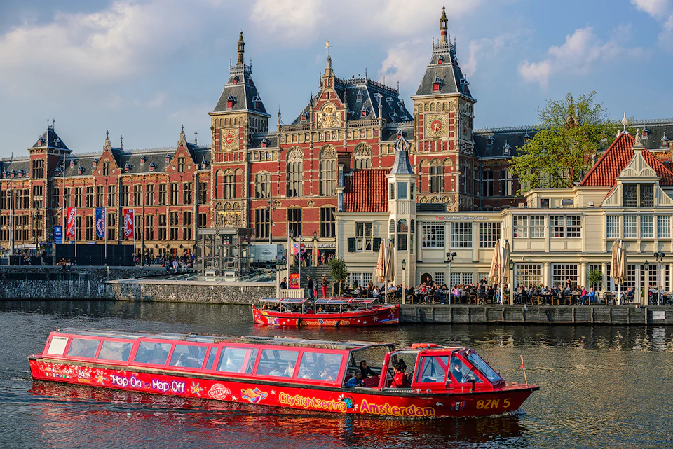 A red sightseeing boat on a canal with a historic brick building in the background, labeled "City Sightseeing Amsterdam."