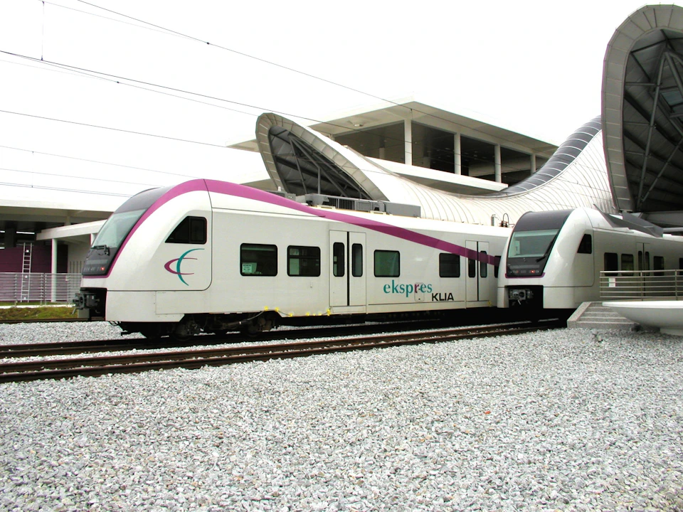 Modern white train with a magenta stripe at a station, labeled "ekspres KLIA," next to a large, curved structure. Gravel-covered tracks.