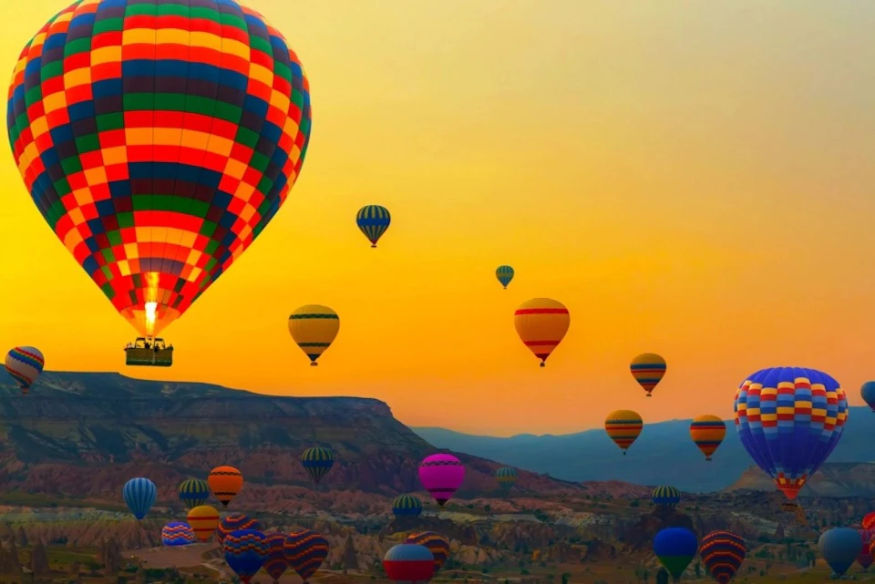 Numerous colorful hot air balloons float over a rugged landscape at sunset.