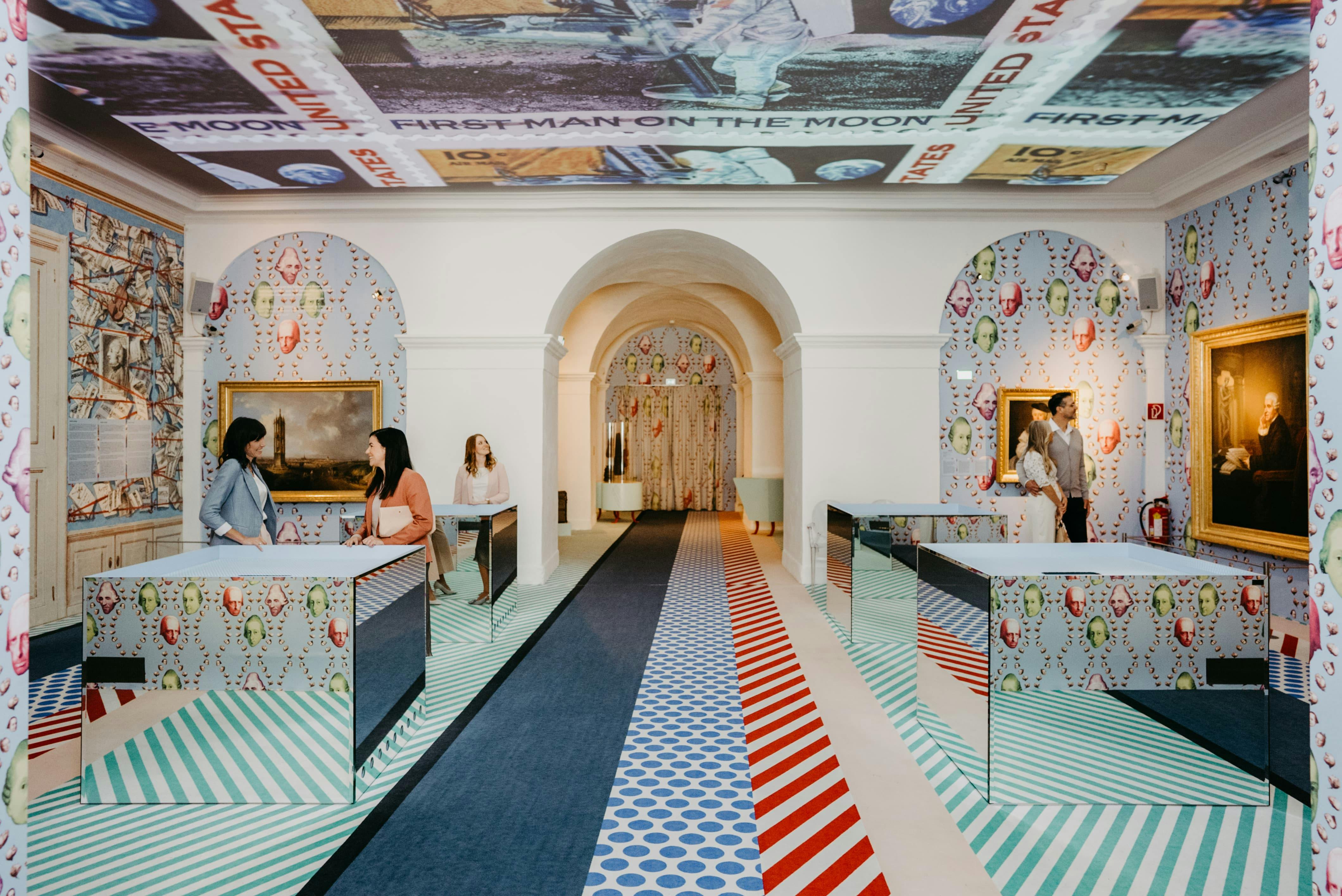 Δύο γυναίκες συνομιλούν πάνω από θρανία με καθρέφτες σε ένα ζωντανό δωμάτιο με σχέδια που διαθέτει καμάρες, έναν πίνακα ζωγραφικής και μια τοιχογραφία οροφής αστροναύτη.