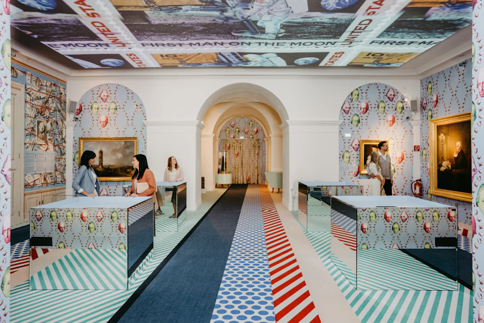 Two women converse over mirrored desks in a vibrant, patterned room featuring archways, a painting, and an astronaut ceiling mural.