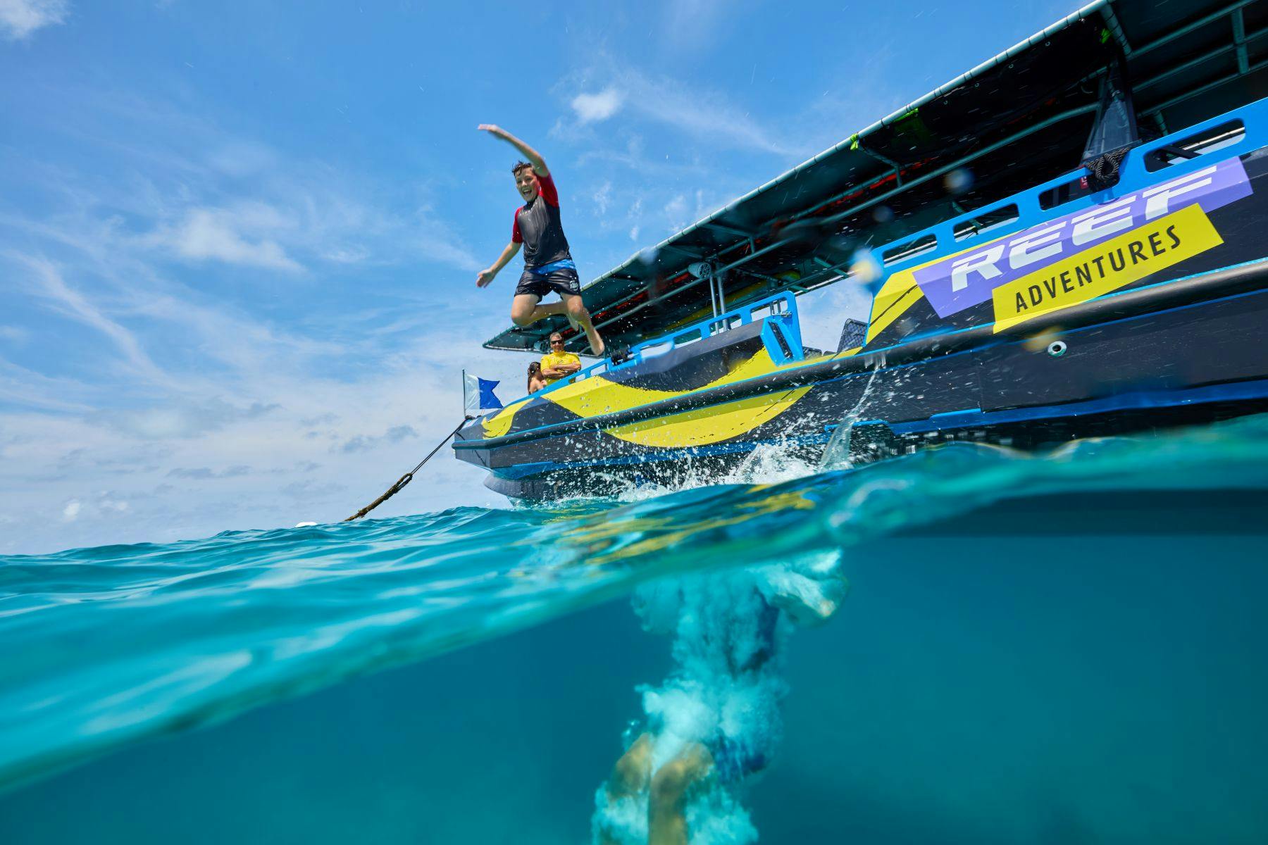 カラフルなグラフィックを持った人物が澄んだ青い海に飛び降り、もう一人が水中を泳いでいる。