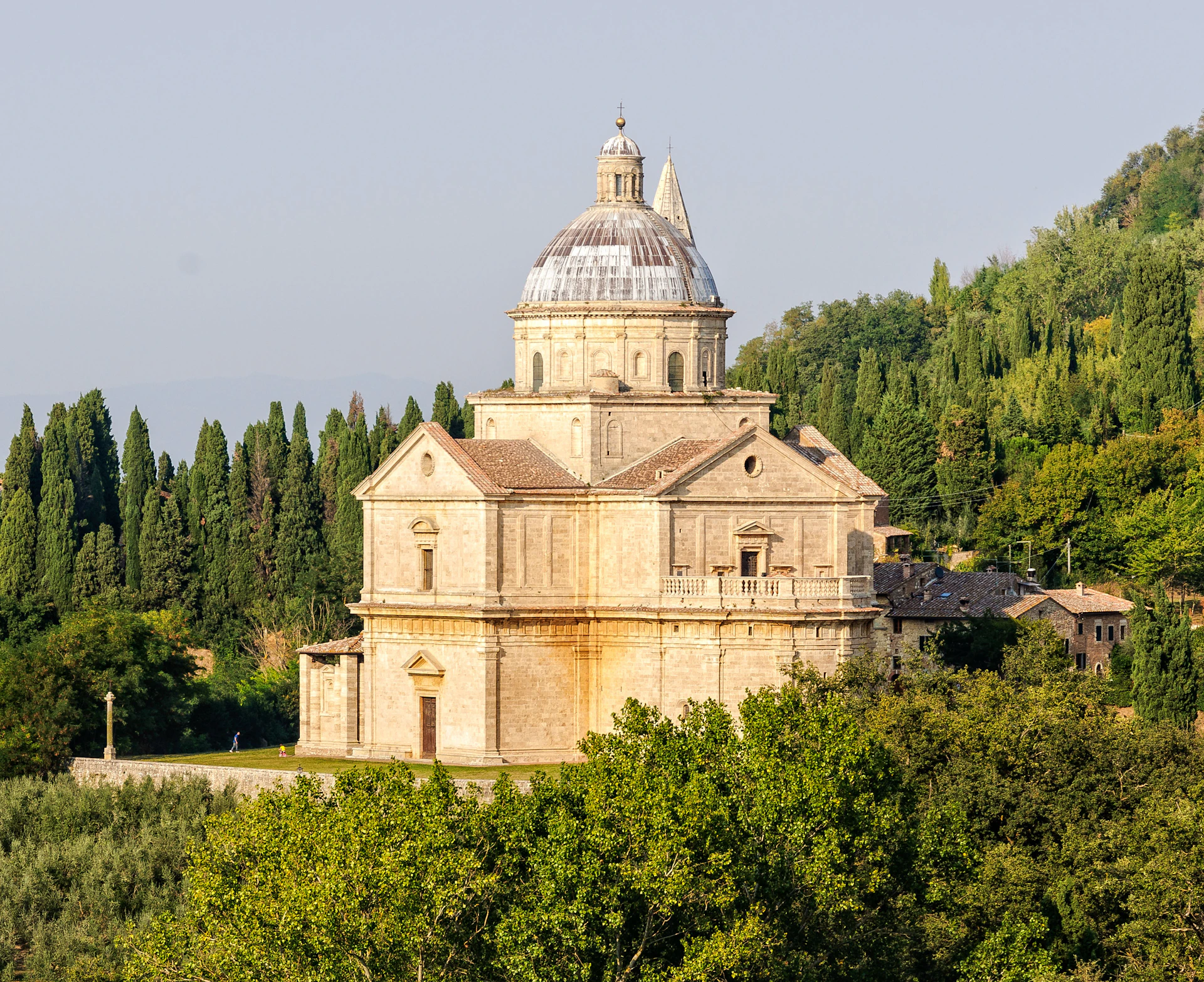 An old, domed stone building with classical architecture is surrounded by trees on a sunny day.