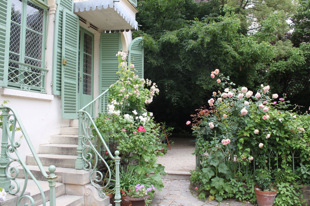 En trädgård med rosor och grönska framför ett hus med gröna fönsterluckor och en liten trappa.