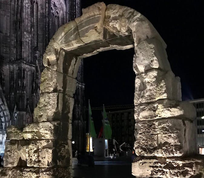 Μια αρχαία πέτρινη αψίδα που φωτίζεται τη νύχτα, με μέρος ενός γοτθικού καθεδρικού ναού ορατό στο παρασκήνιο.