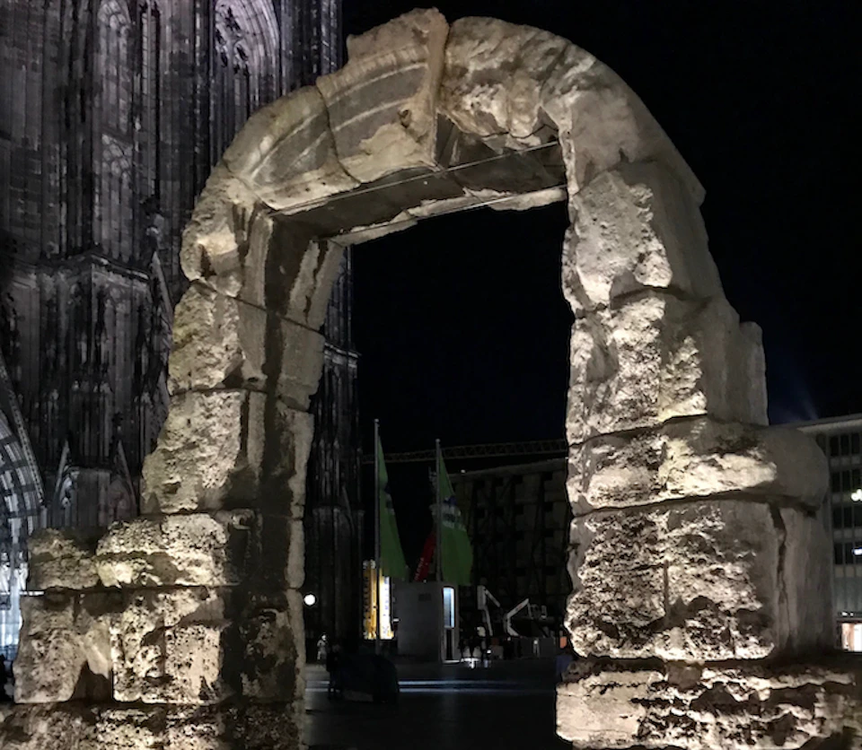 An ancient stone archway illuminated at night, with part of a gothic cathedral visible in the background.