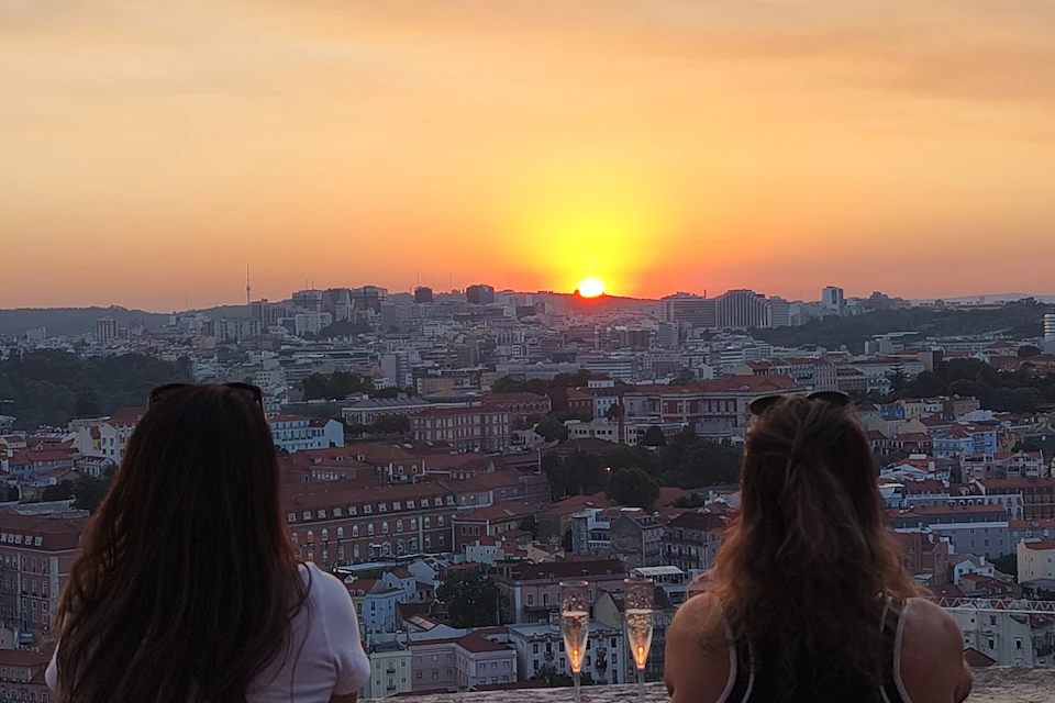 Two people facing a cityscape during sunset, with two glasses of champagne on the ledge in front of them.