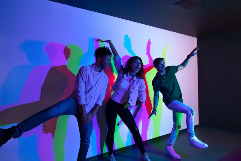 Three people pose playfully in front of a white wall with colorful shadow projections.