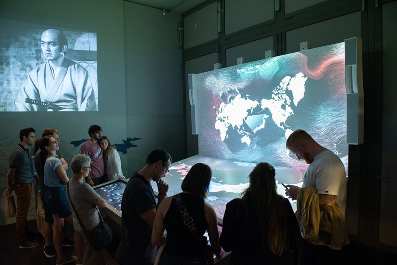 La gent observa una taula interactiva en una habitació poc il·luminada, amb una projecció de mapa i una pel·lícula en blanc i negre a la paret.