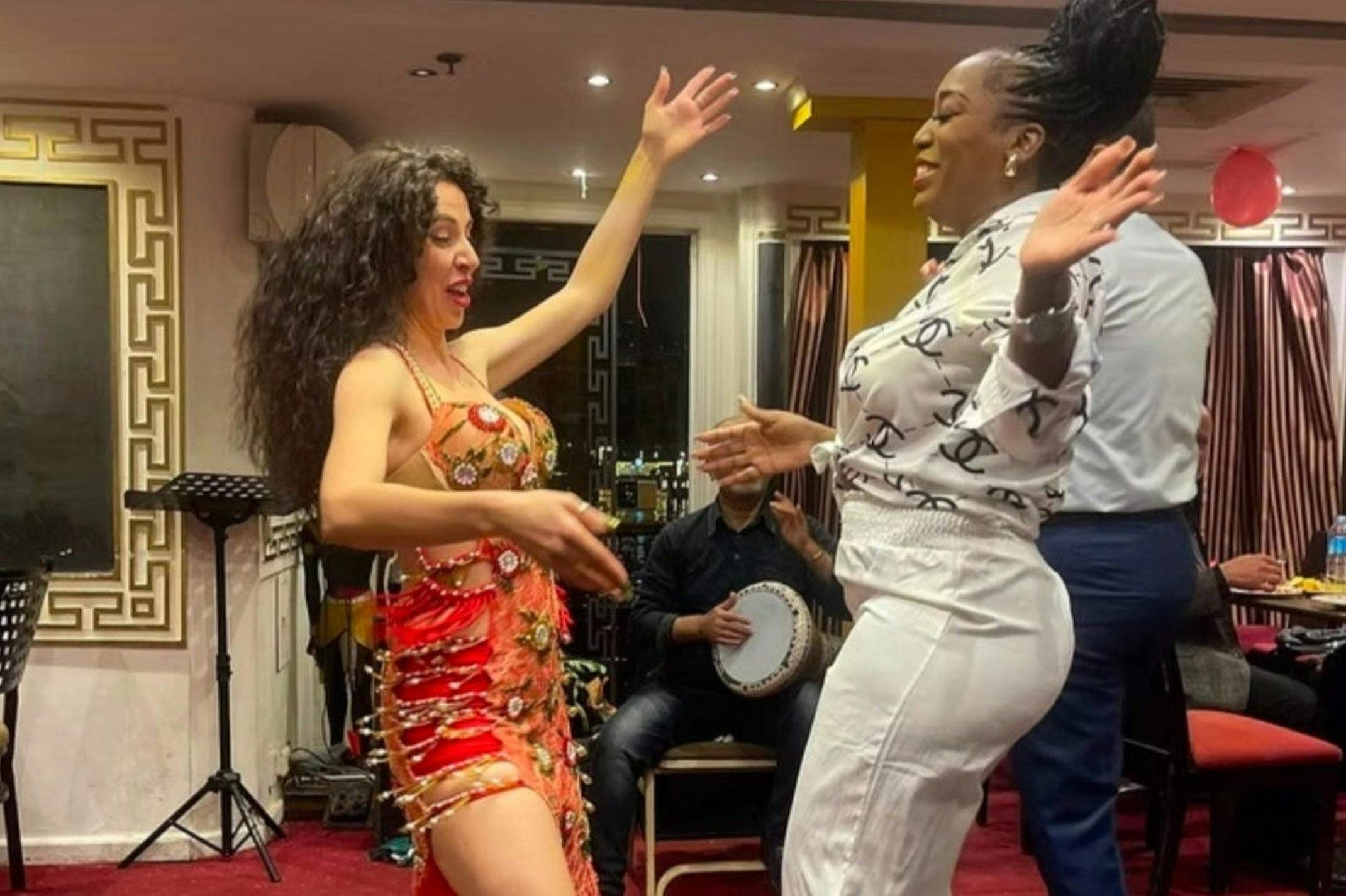 Zwei Frauen tanzen fröhlich, während ein sitzender Musiker im Hintergrund eine Trommel spielt, in einem Innenraum mit warmer Beleuchtung.