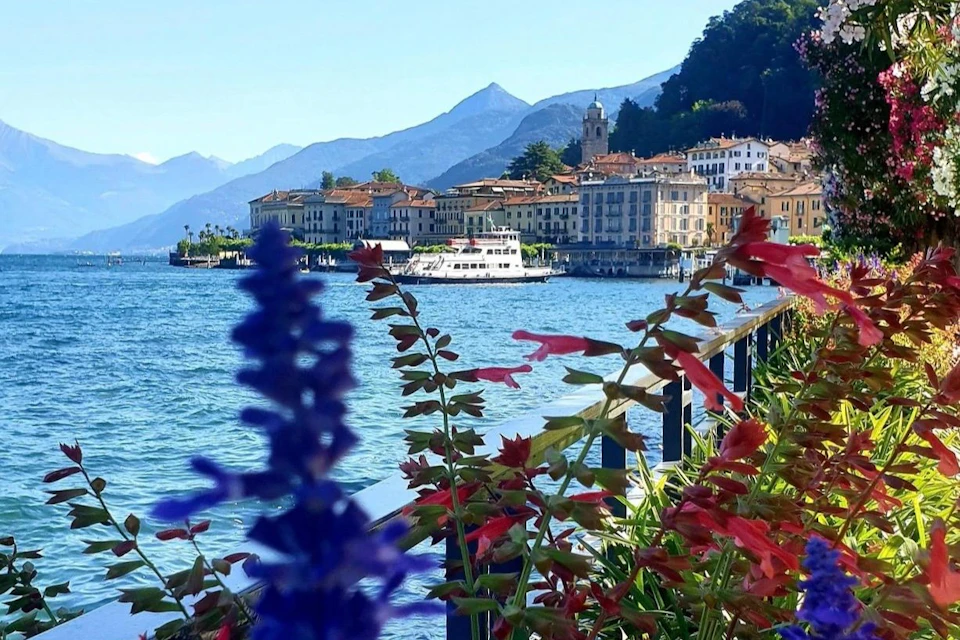 Colorful flowers frame a view of a lakeside town with a docked boat, surrounded by mountains under a clear blue sky.