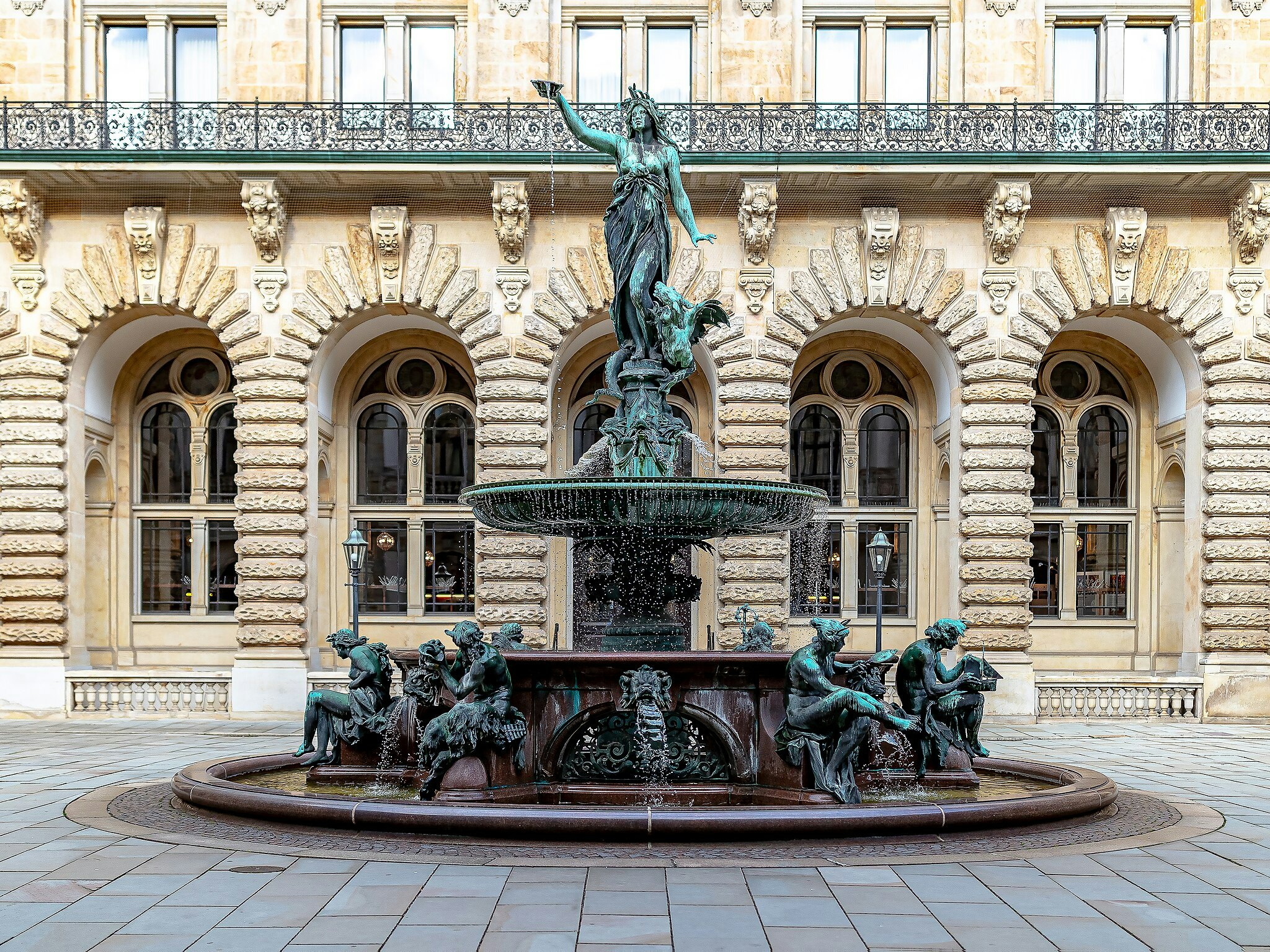 Fonte com estátua de bronze em frente a um edifício histórico com janelas em arco, cantarias pormenorizadas e uma balaustrada.