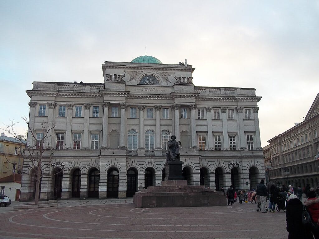 Edificio neoclásico ornamentado con tejado abovedado, ventanas arqueadas y una estatua de una figura sentada en primer plano, gente paseando cerca.