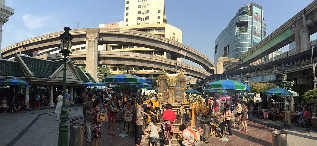 在一个繁忙的市区，人们聚集在一个蓝色雨伞下的小神社周围，背景是高架混凝土高速公路和建筑物。