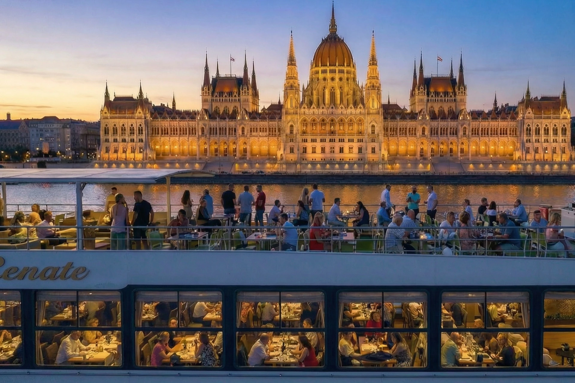 A boat with dining passengers on a river, with a lit historic building and a sunset sky in the background.