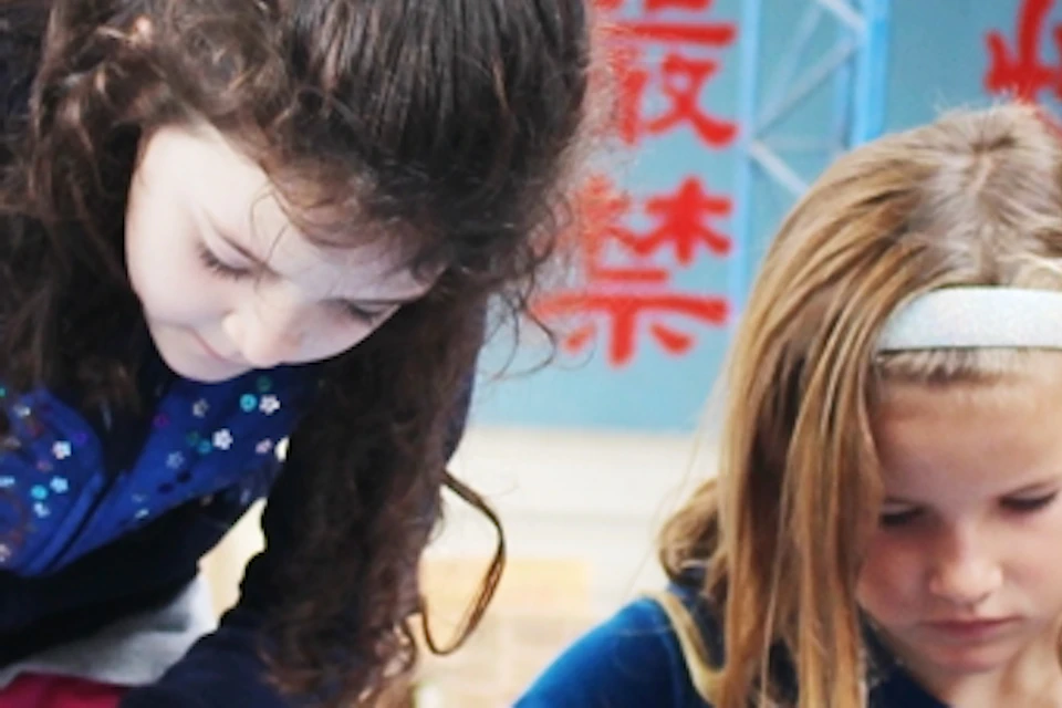 Children playing with colorful objects in a blue box in a covered outdoor area with visible beams and a roof.
