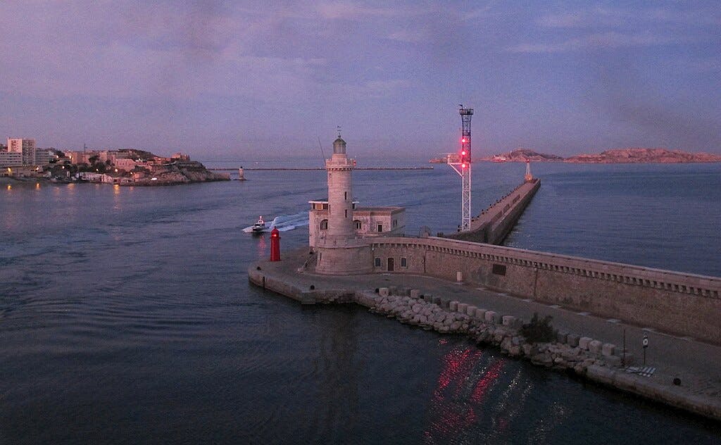 Маяк и небольшая красная башня на пирсе, когда проплывает лодка; прибрежные здания и острова на заднем плане в сумерках.