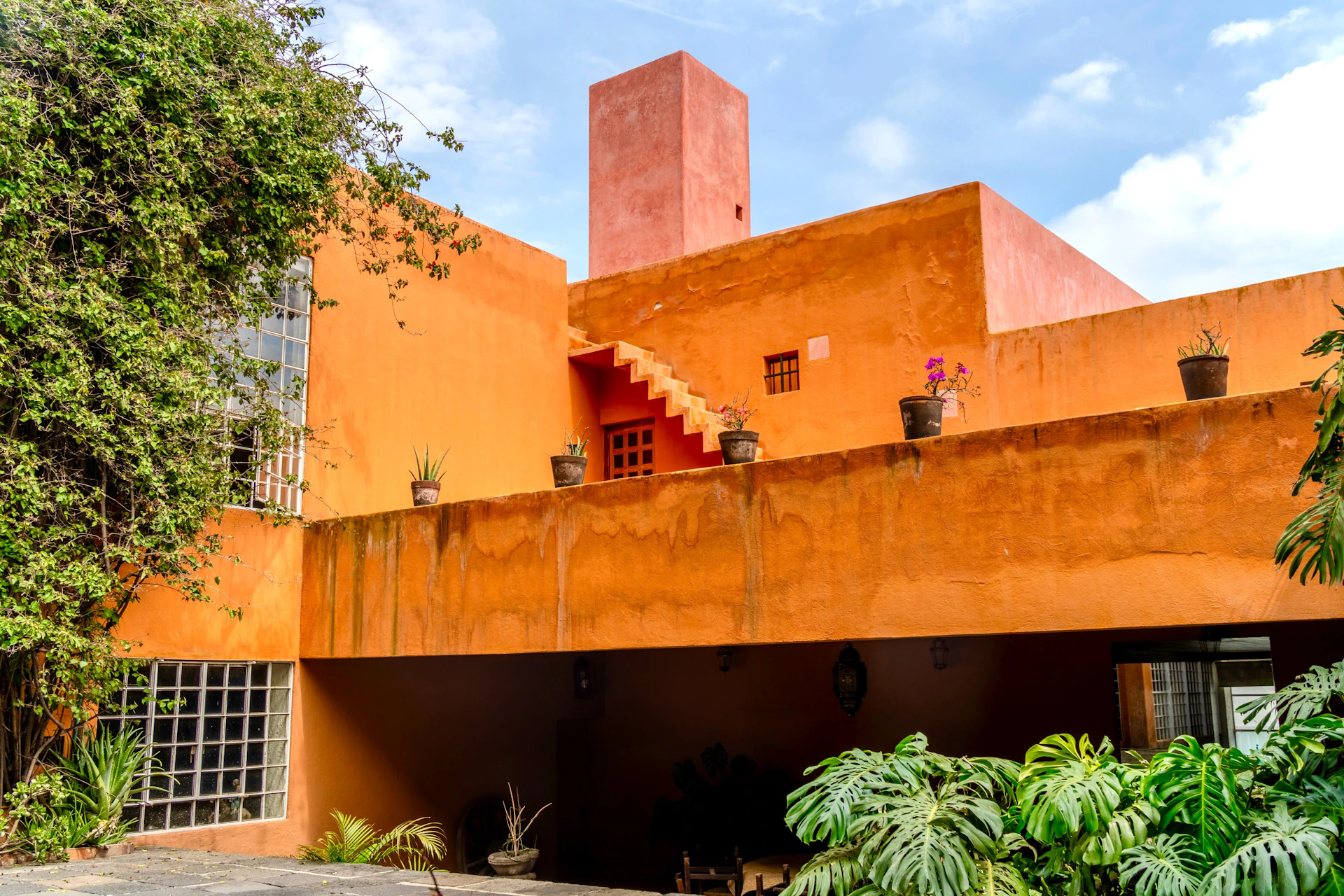 A bright orange building with a rooftop terrace, potted plants, and a staircase. Large green foliage is in the foreground.