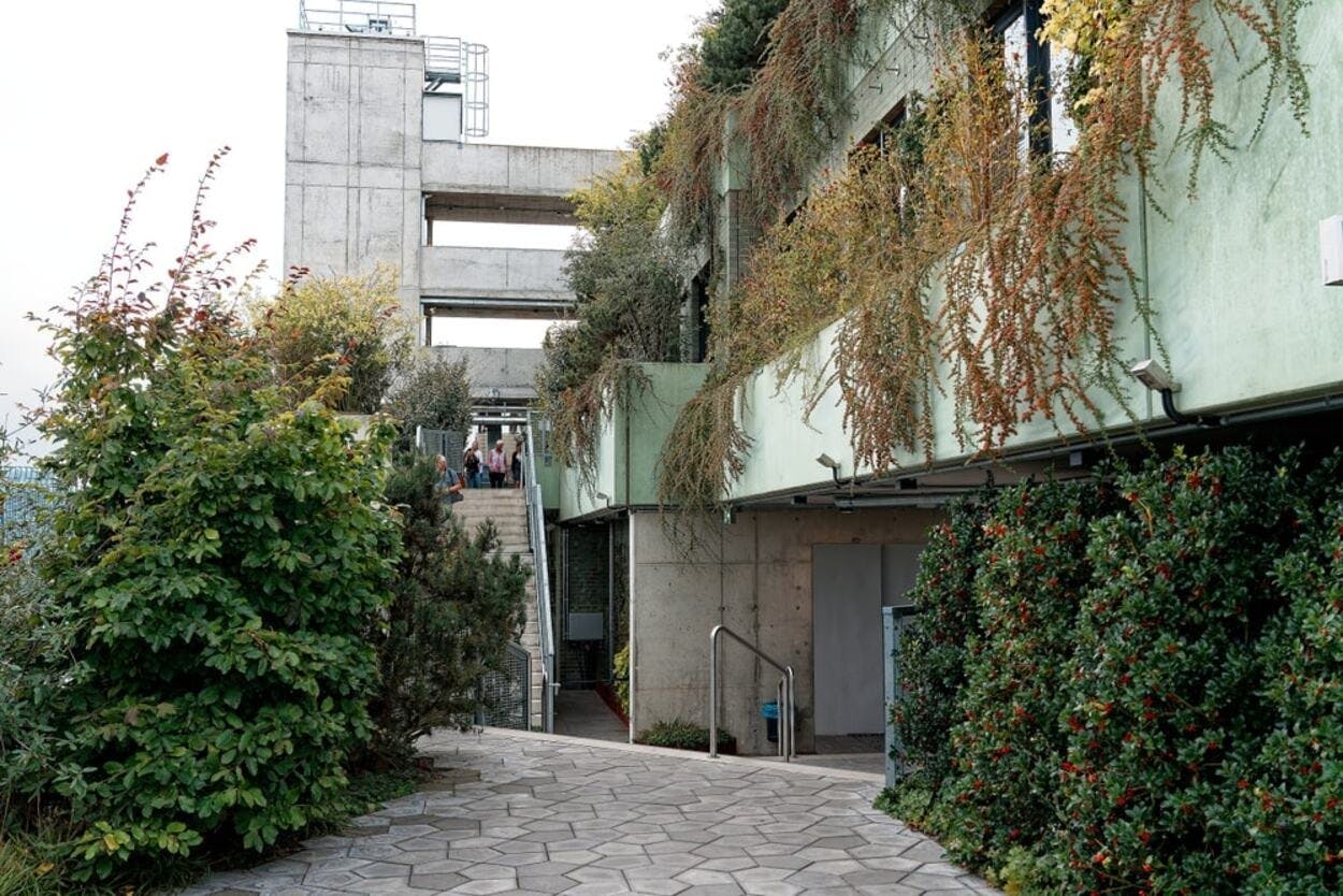 Betonová budova se zelenými stěnami pokrytými rostlinami, schodištěm a cestami obklopenými bujnou zelení. V dálce jsou vidět lidé.