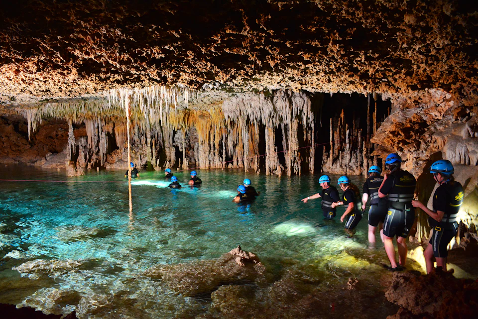 People wearing helmets and wetsuits explore a cave with stalactites and clear blue water.