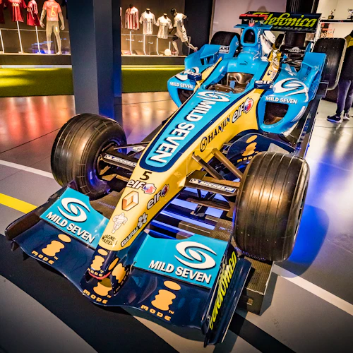 Ein blau-gelber Rennwagen, der in einem Museum ausgestellt ist, mit zwei Personen, die sich die Exponate im Hintergrund ansehen.