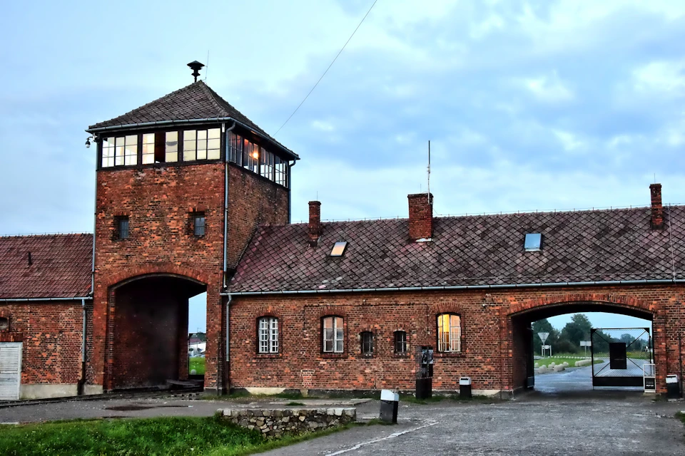 A brick building with an arched gate and a tower, set under a cloudy sky with a grassy area and path in front.